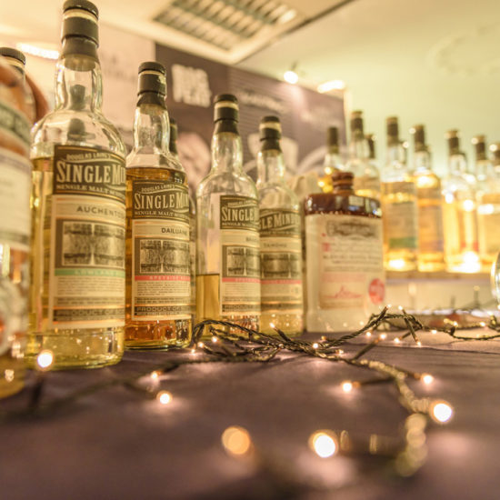 Eine Reihe fast gleicher Single Malt Whiskyfalschen steht auf einem Tisch mit Lichterkette und einem Glaskrug mit Wasser.