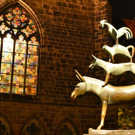 Die Bronzeskulptur der Bremer Stadtmusikanten vor einem erleuchten Kirchenfenster.