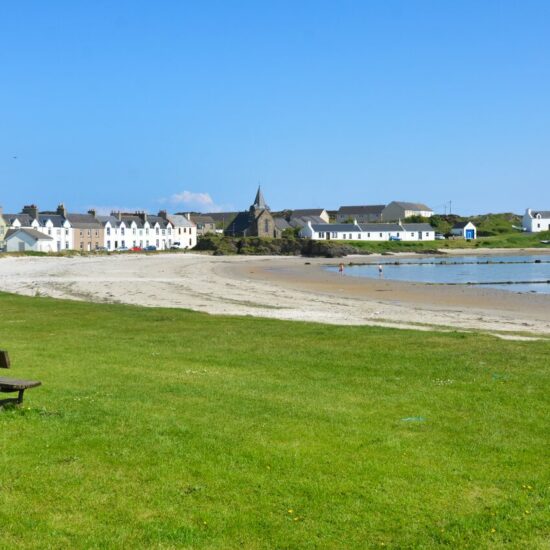 Blick über das Hafenbecken von Port Ellen auf Islay, auf der Wiese steht eine Bank und im Hintergrund liegt eine Reihe kleiner weißer Wohnhäuser.